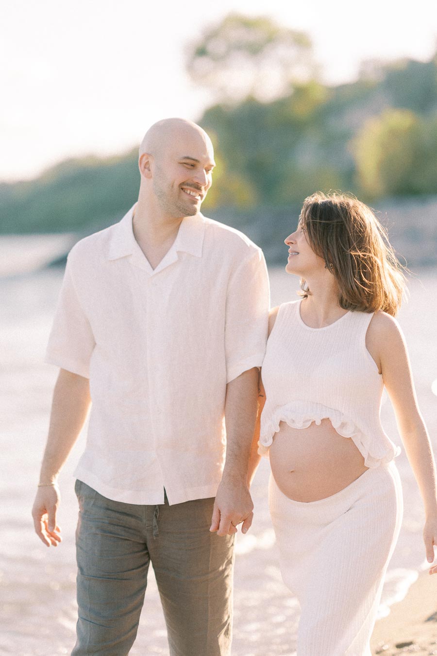 Expectant couple walking hand in hand along a sunny beach shoreline.