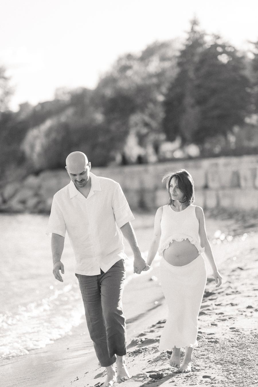 Pregnant woman and partner hold hands while walking barefoot along a sandy beach.
