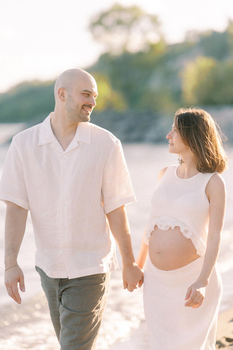 Expectant couple holding hands and walking along a sunlit beach near the water.
