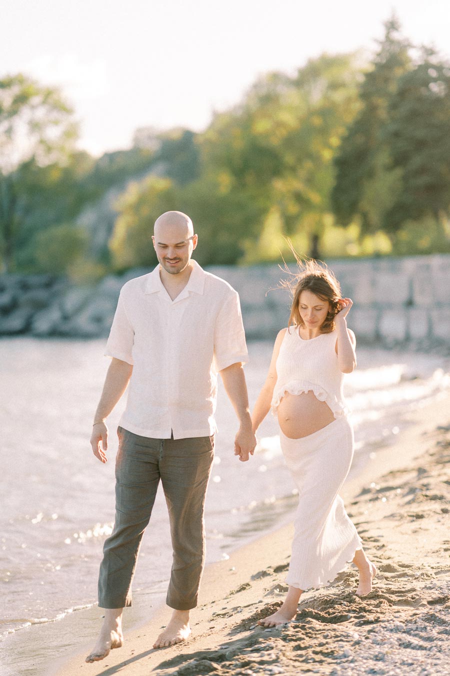 Expectant couple walking barefoot hand in hand along a sandy beach by the water.
