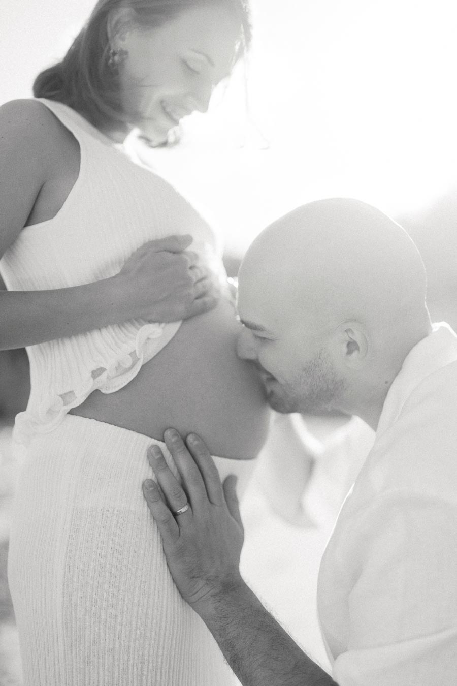 A man kisses a pregnant woman's belly while she smiles in soft backlight.