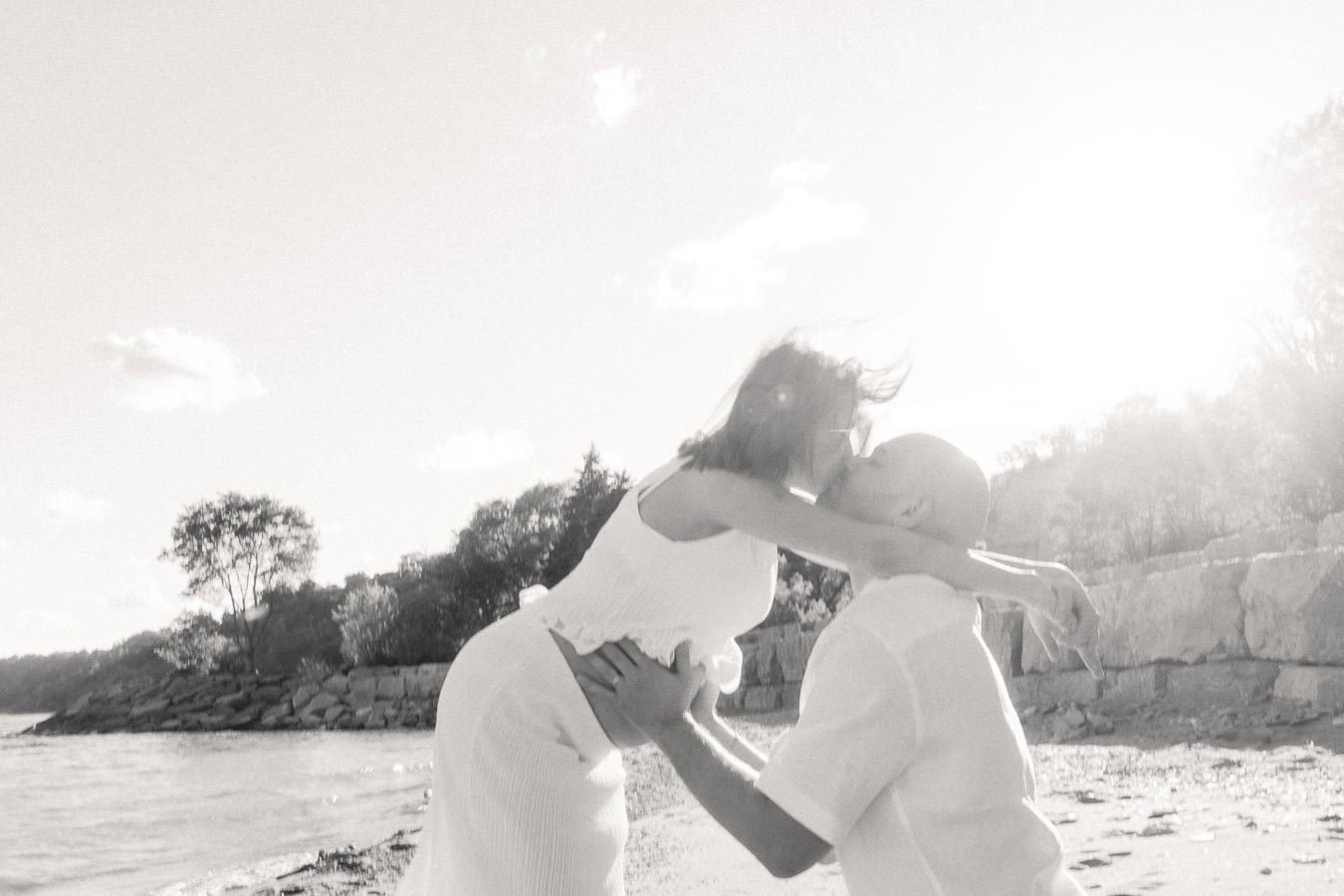 Pregnant woman embraces a kneeling man on a sunlit beach beside rocky bluffs.