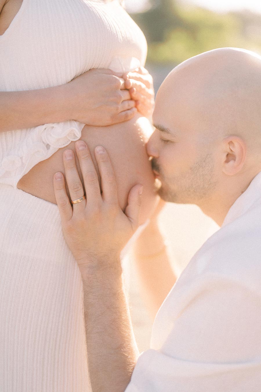 Expectant father kisses pregnant belly while both parents hold it in warm sunlight.