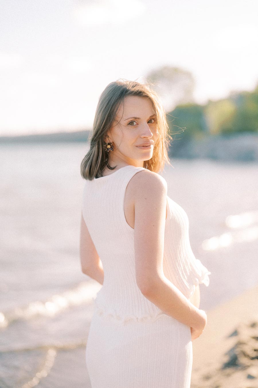 Pregnant woman in a white dress looks back on a sunlit beach beside the water.
