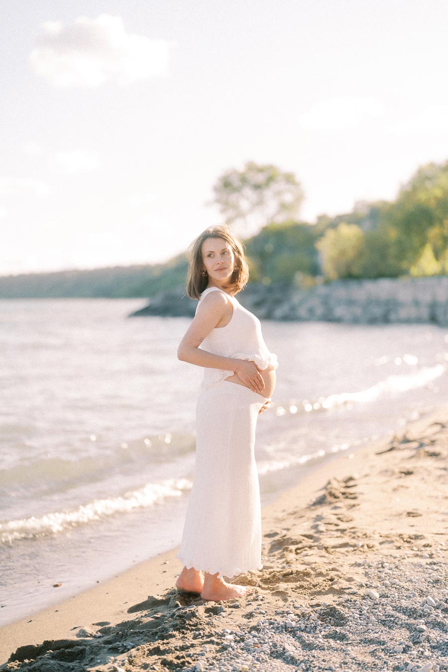 Pregnant woman in a white dress standing barefoot on a sandy beach beside the water.