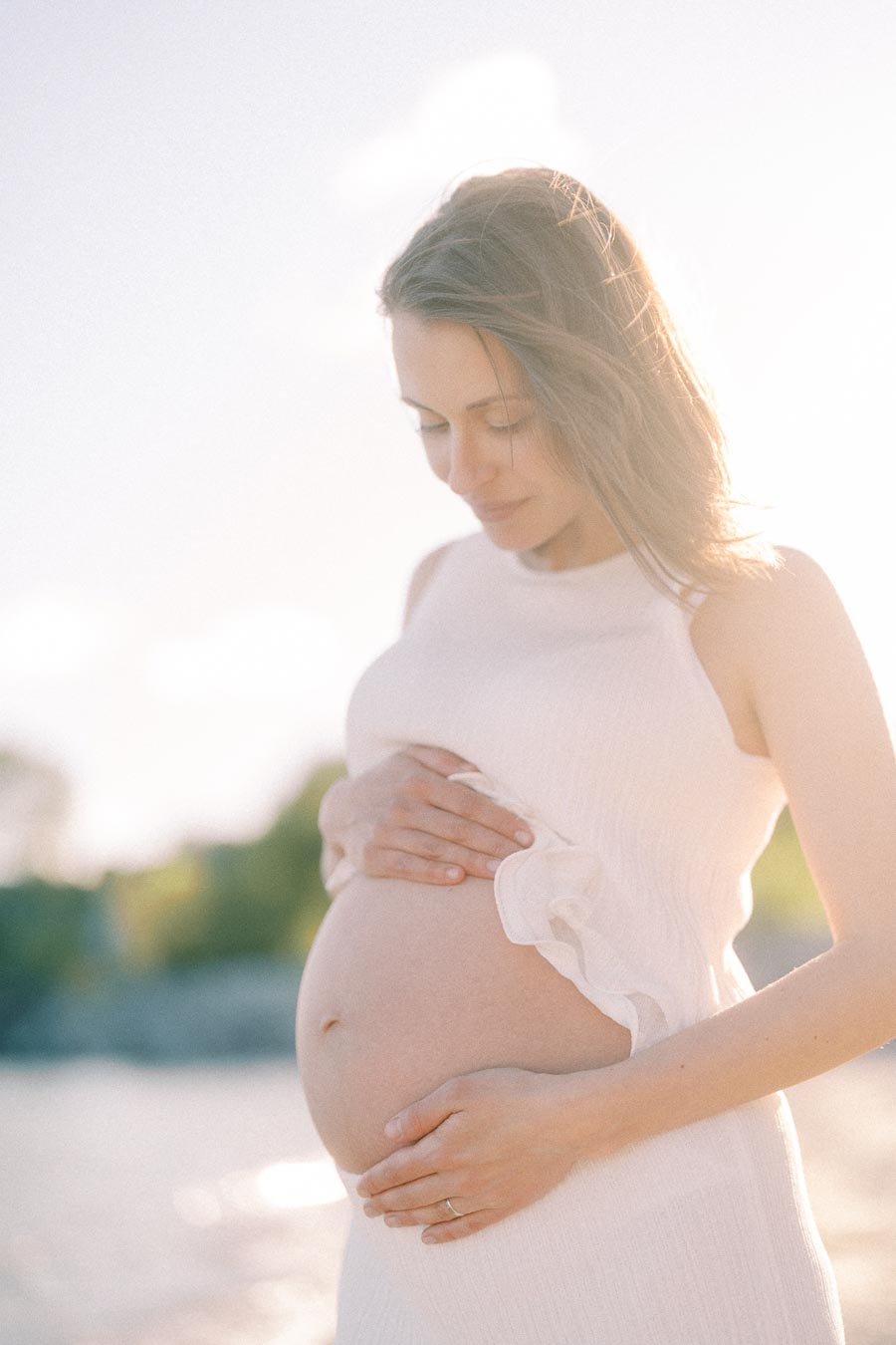 Pregnant woman in a white dress gently holds her belly in soft backlight.