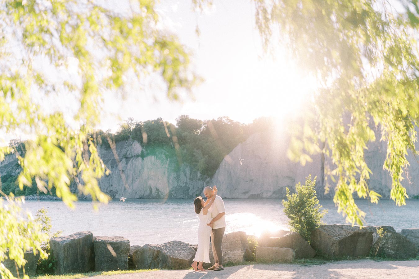 Couple embracing by Lake Ontario with Scarborough Bluffs cliffs behind them in bright sunlight.