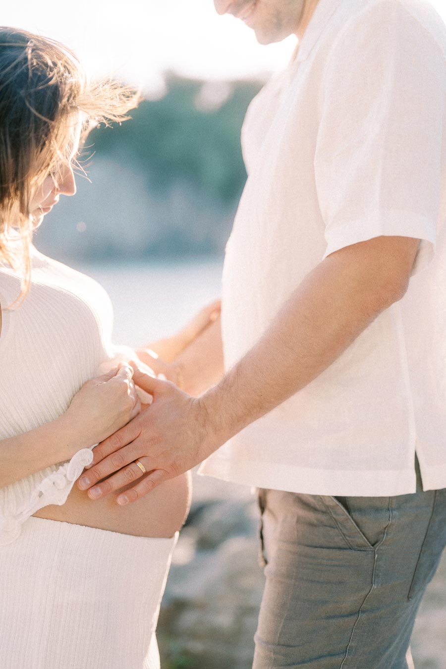 Expectant parents gently hold a pregnant belly in soft backlight outdoors.