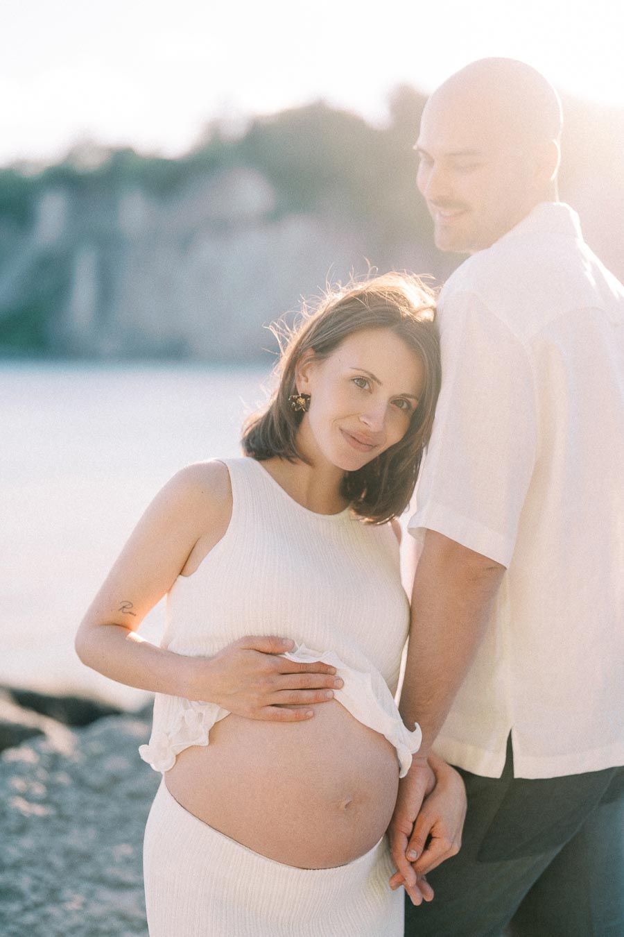Pregnant woman in white leans beside her partner near the water at Scarborough Bluffs.