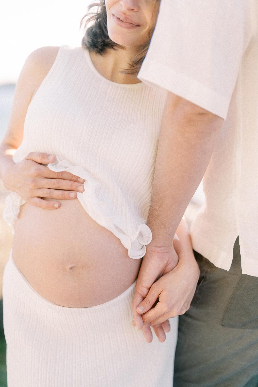 Pregnant woman in white holds hands with her partner beside her baby bump.