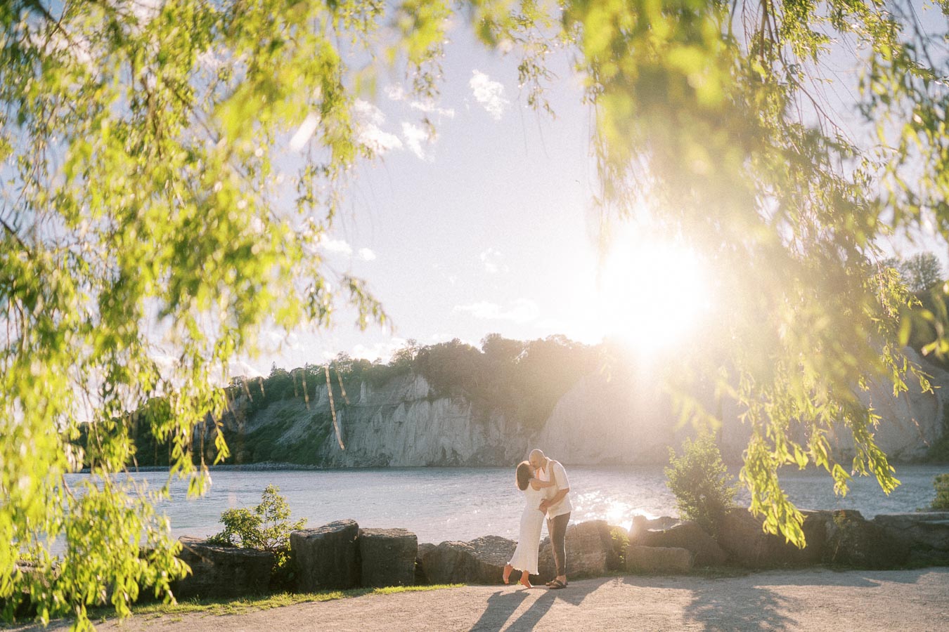 Expectant couple kissing by Lake Ontario with Scarborough Bluffs and sunlit trees behind them.