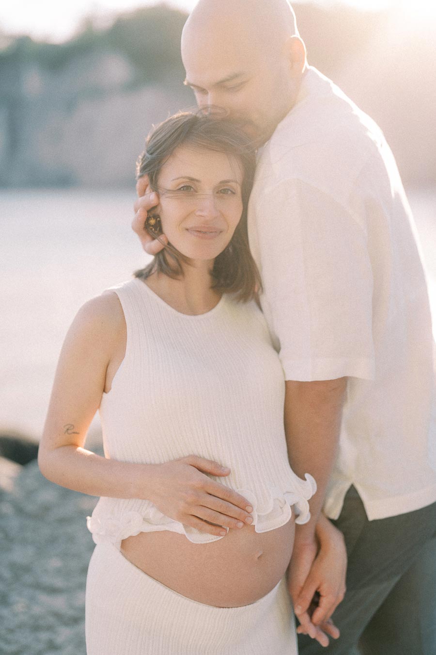 Expectant couple embracing by the water, with the woman’s pregnant belly visible in soft sunlight.