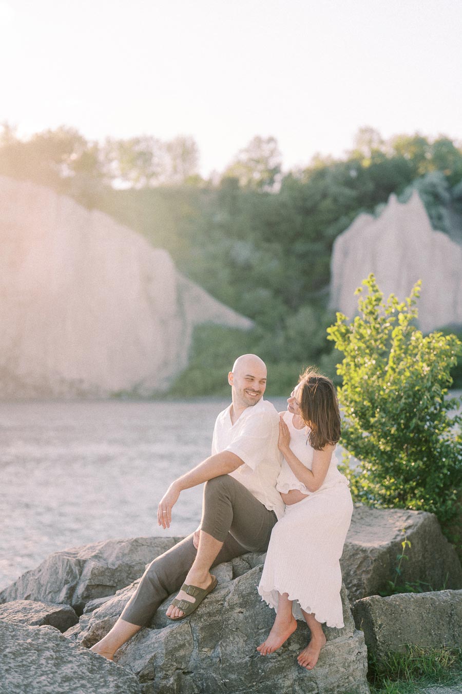 Pregnant woman in a white dress sits with her partner on rocks at Scarborough Bluffs.