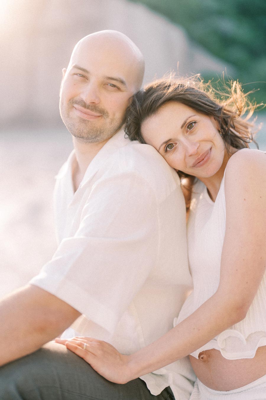Pregnant woman rests her head on her partner’s shoulder in warm outdoor light.