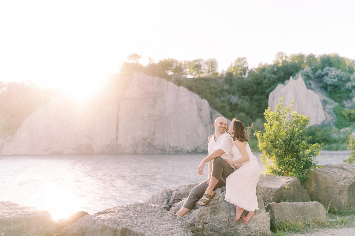 Expectant couple sitting on rocks beside Lake Ontario with sunlit Scarborough Bluffs behind them.