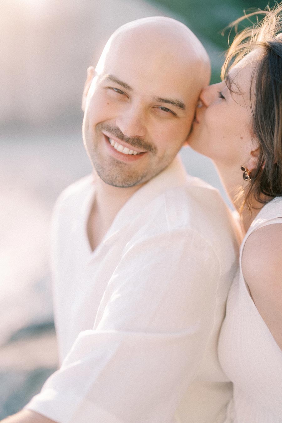 Pregnant woman kisses smiling partner on the temple in soft outdoor light.