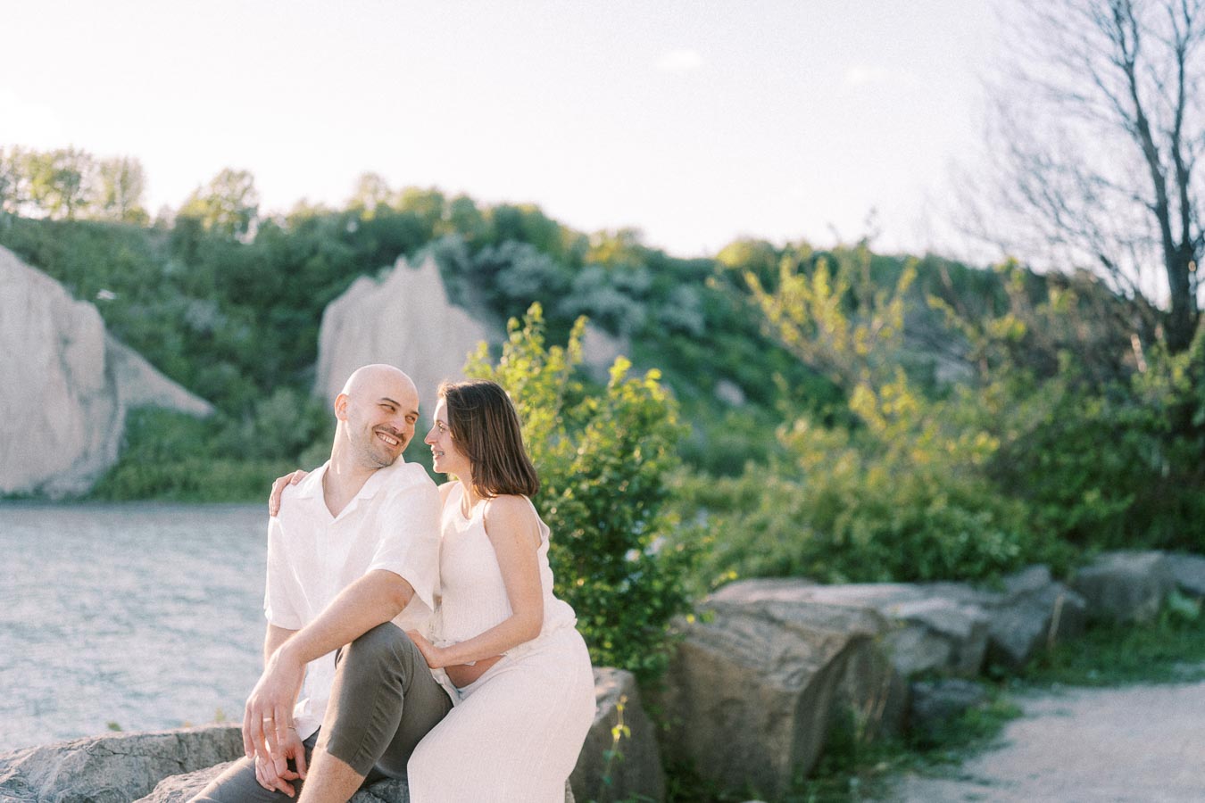 Expectant couple sitting together on rocks beside water and cliffs at Scarborough Bluffs.