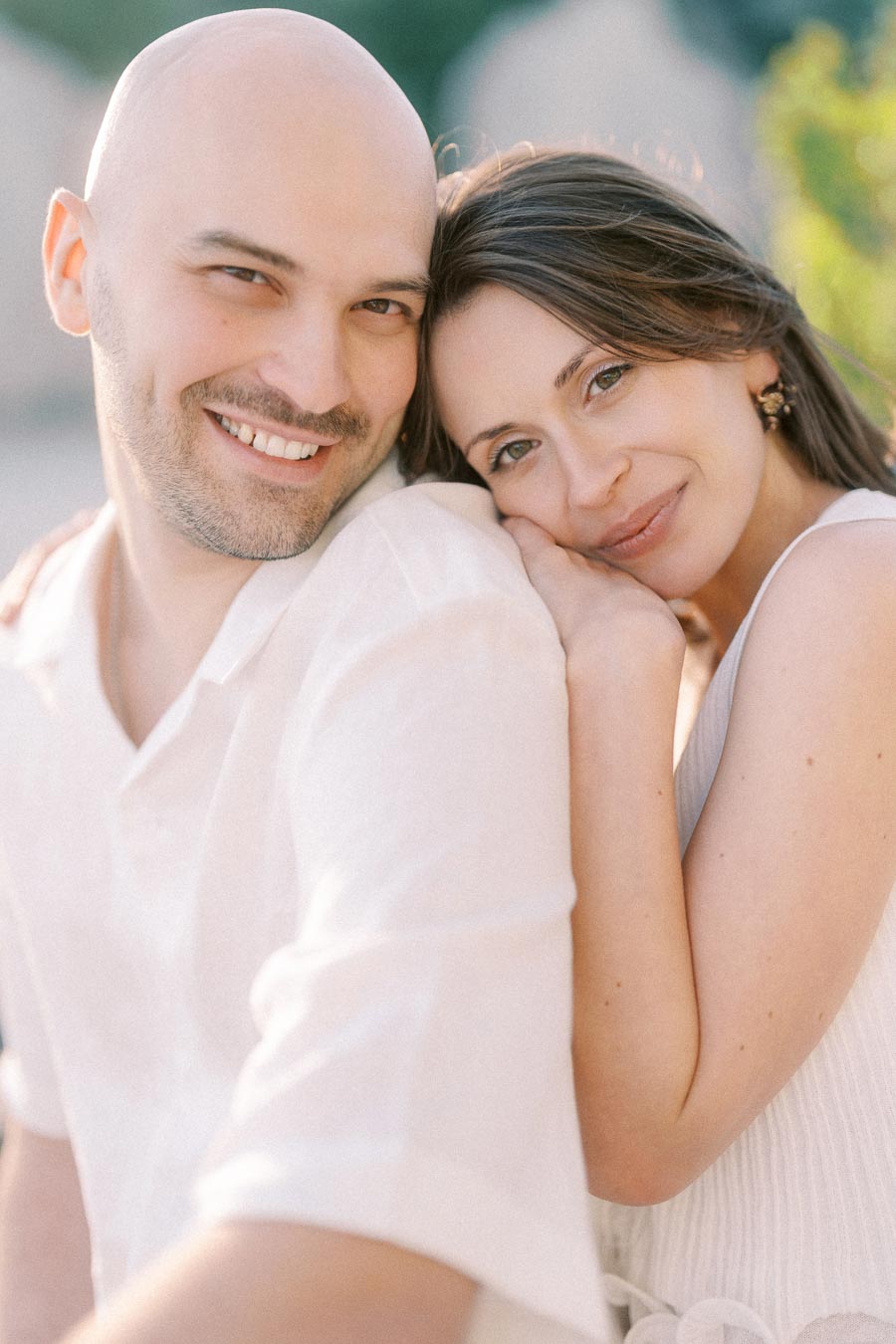 Close-up portrait of a smiling couple in white clothing, with the woman resting on his shoulder.