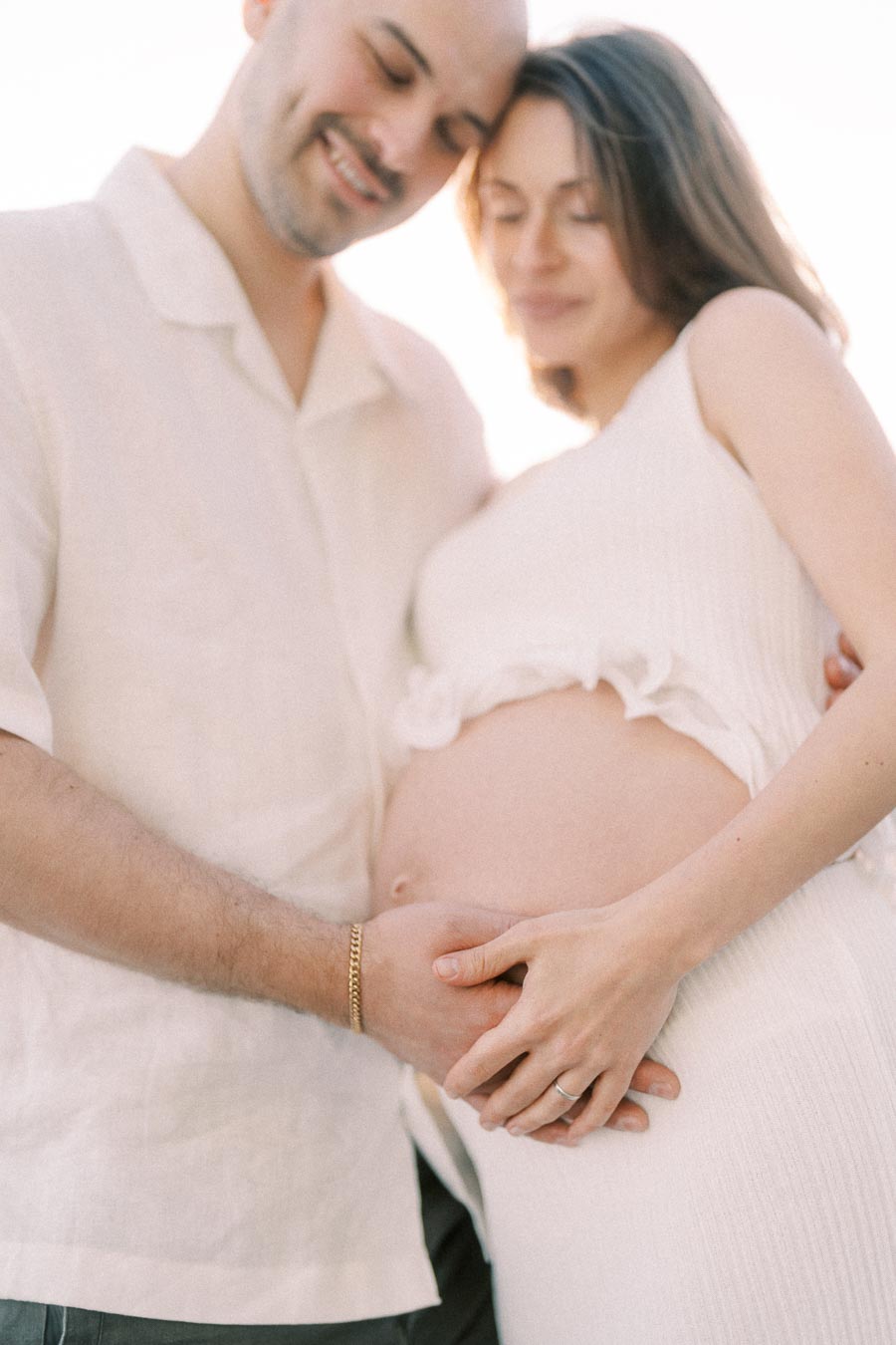 Expectant couple in white clothing holding hands over the pregnant belly in soft sunlight.