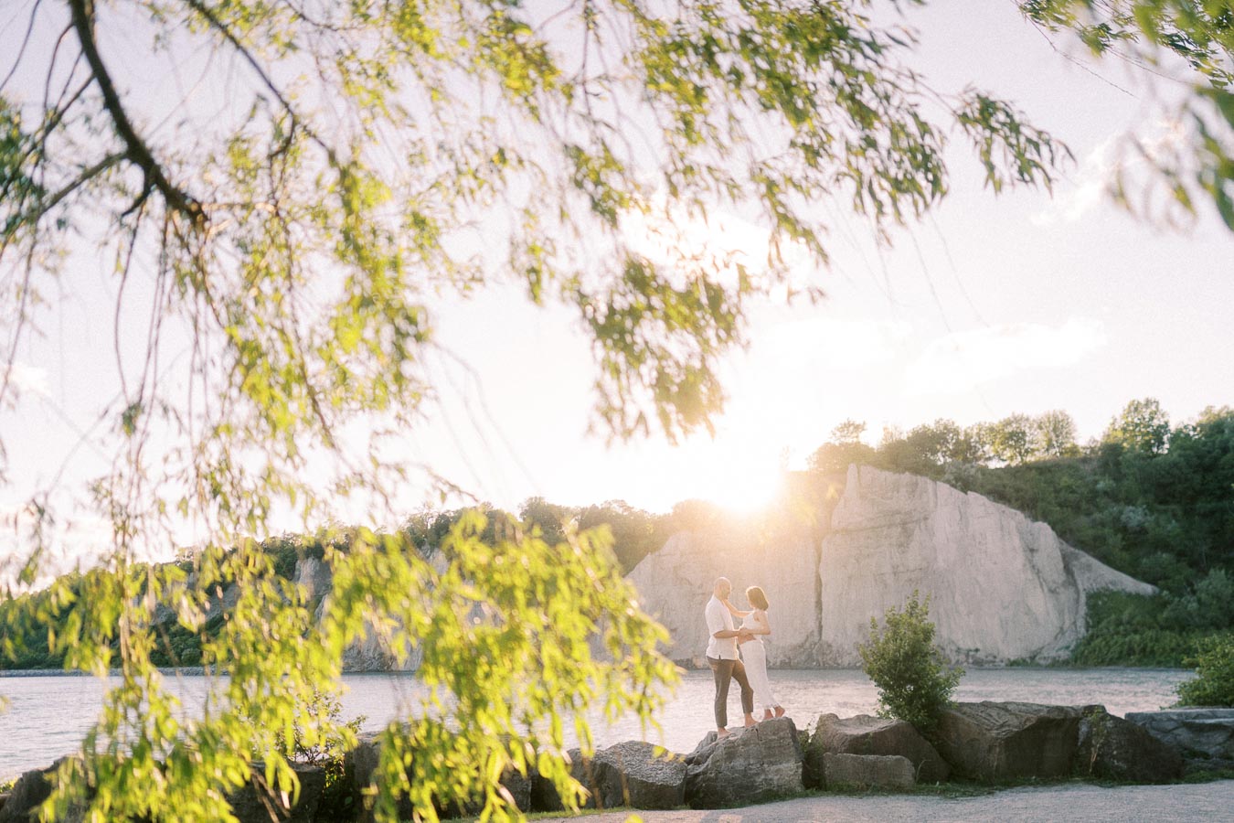 Expectant couple stands on lakeside rocks beneath tree branches with Scarborough Bluffs behind them.