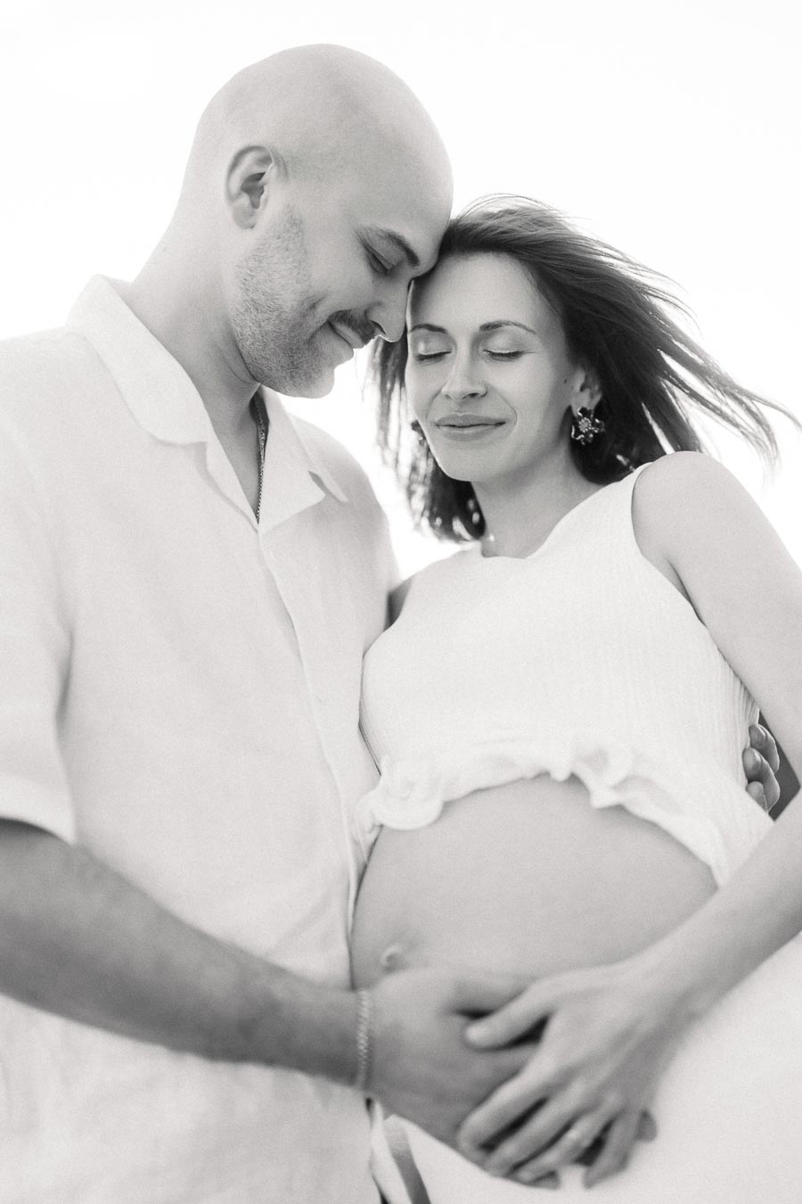 Black-and-white close-up of expectant parents touching the mother’s belly with eyes closed.