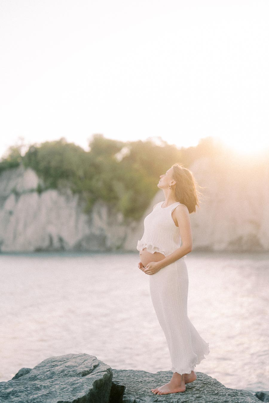 Pregnant woman in a white outfit stands barefoot on rocks by the water at sunset.