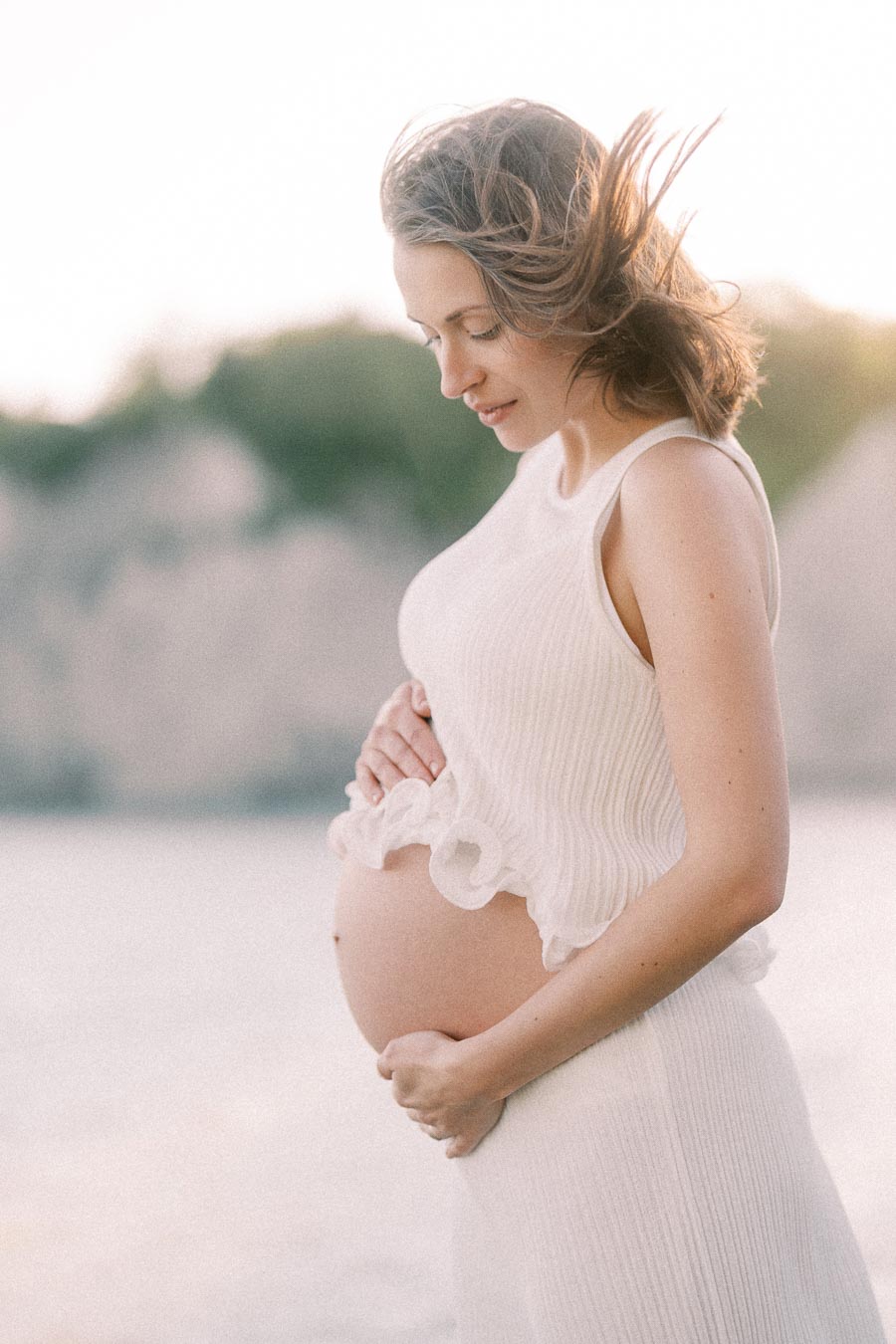 Pregnant woman in a white dress cradles her belly outdoors by softly blurred water.