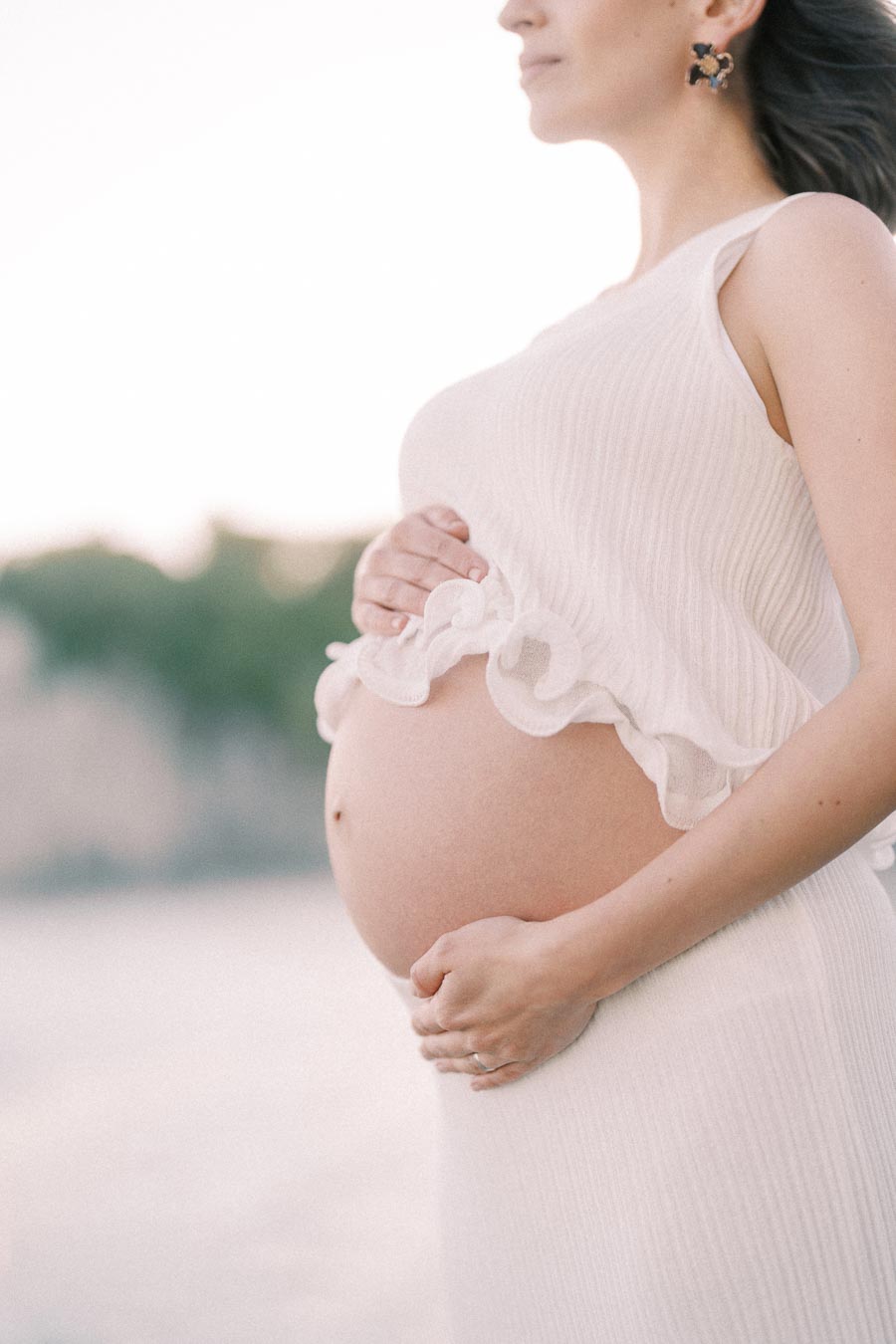 Close-up of a pregnant woman in a white dress holding her belly outdoors.
