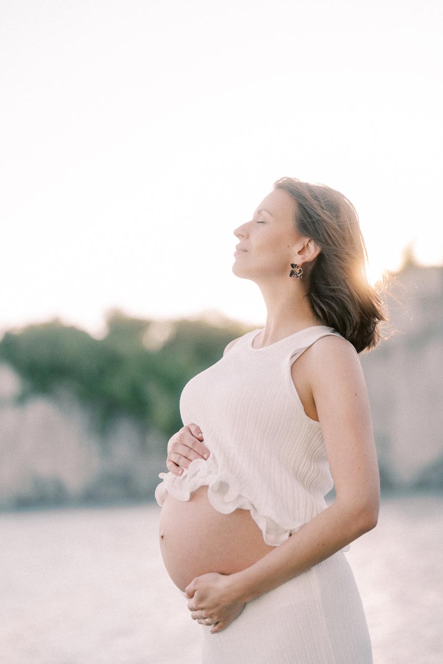 Pregnant woman in a white dress cradles her belly near the water at sunset.