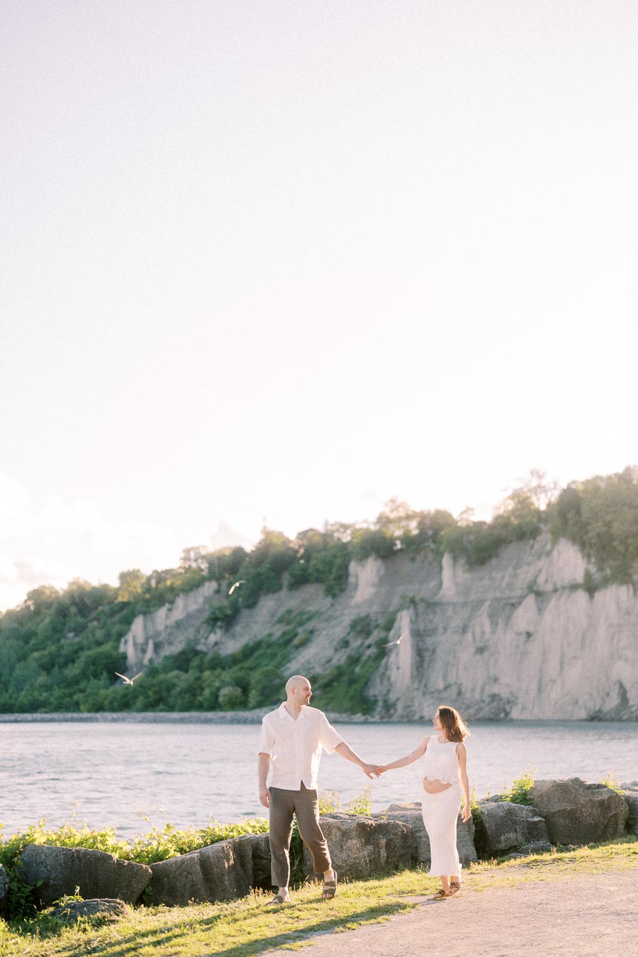 Expectant couple holds hands by Lake Ontario with Scarborough Bluffs in the background.