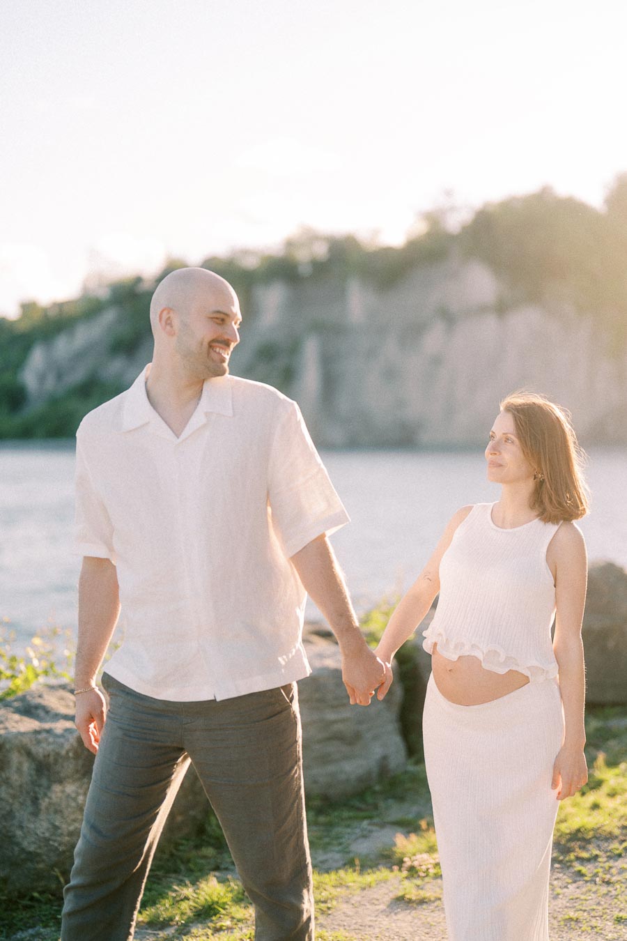 Expectant couple holds hands by Lake Ontario with Scarborough Bluffs in the background.