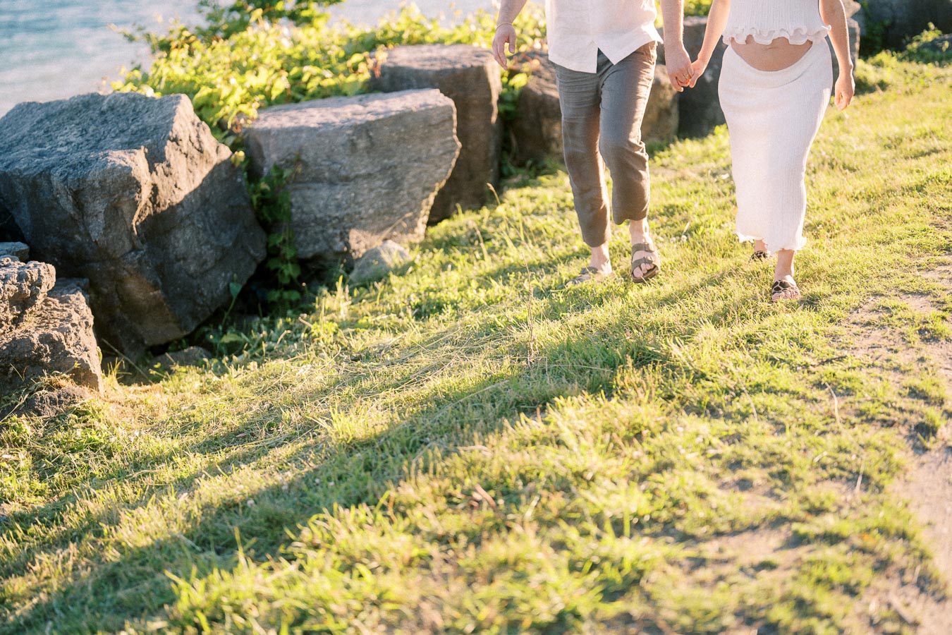 Expectant couple holding hands while walking on a grassy path beside large rocks.