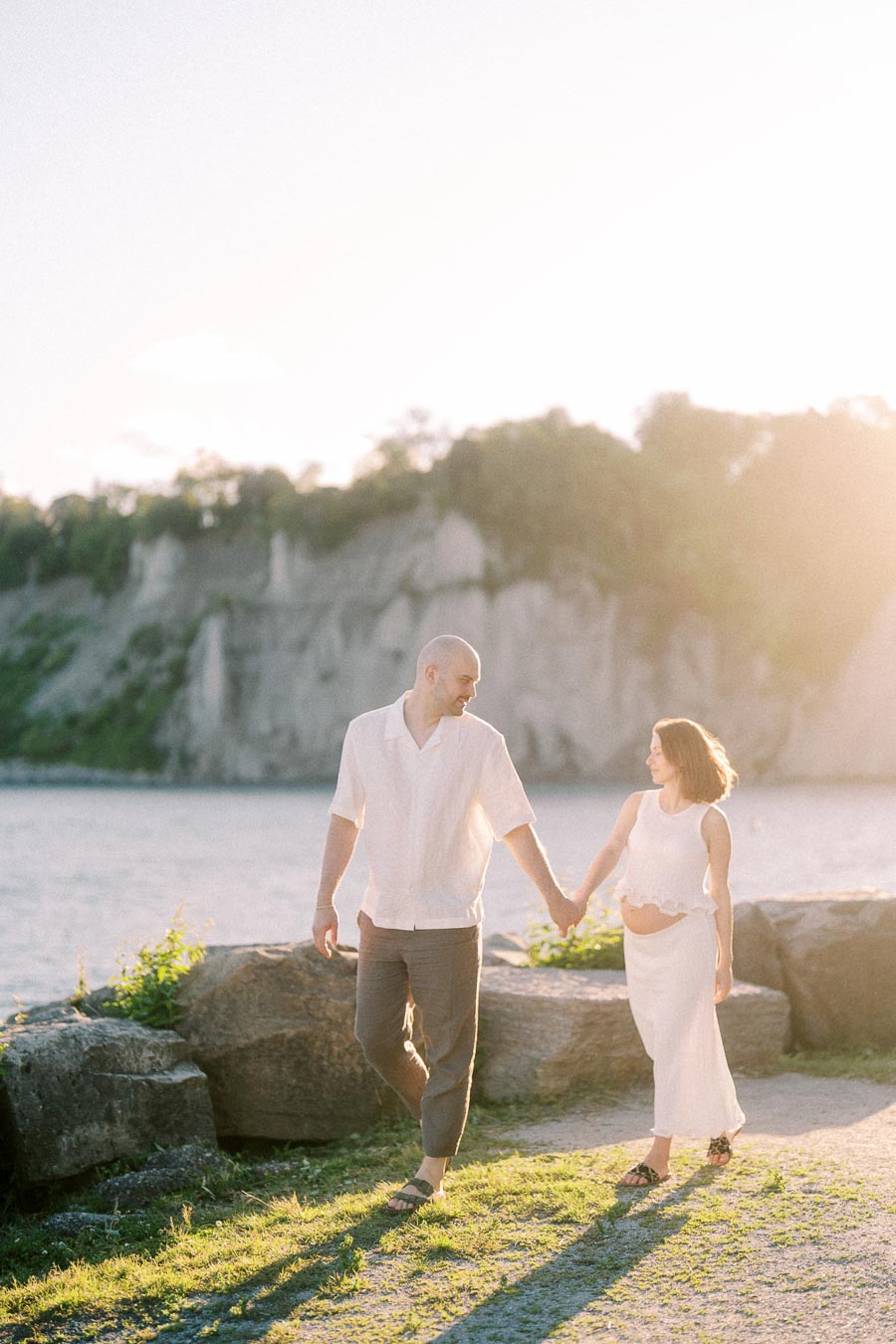 Expectant couple holding hands and walking by Lake Ontario with Scarborough Bluffs in the background.