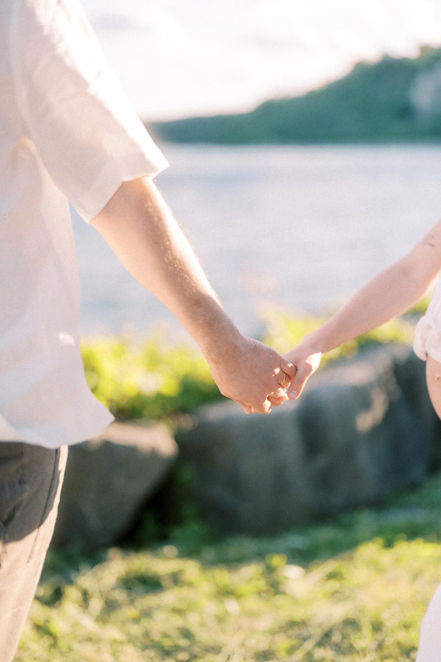 Couple holds hands beside Lake Ontario with sunlit rocks and Scarborough Bluffs in the background.