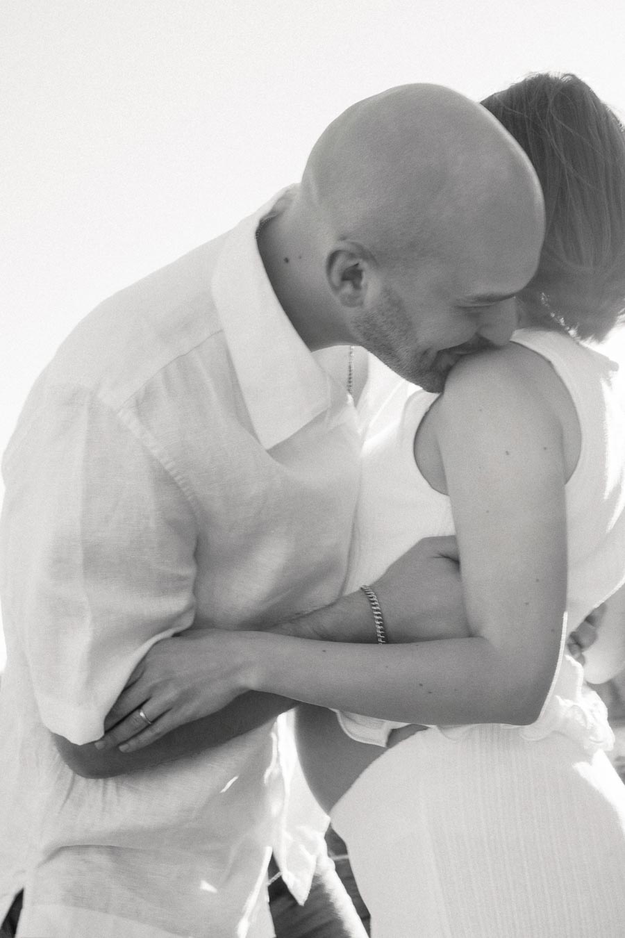 Black and white close-up of an expectant couple embracing, with the man leaning toward her shoulder.