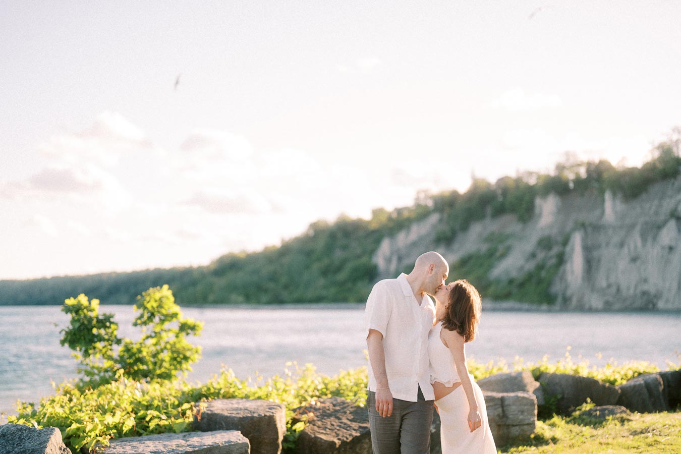 Expectant couple kissing beside Lake Ontario with Scarborough Bluffs cliffs in the background.