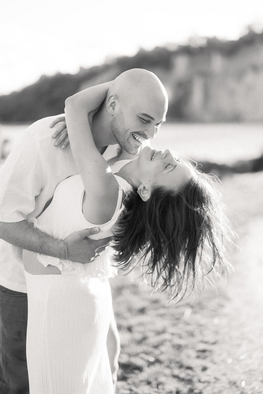 Man dips a pregnant woman in a white dress on a beach with cliffs behind them.