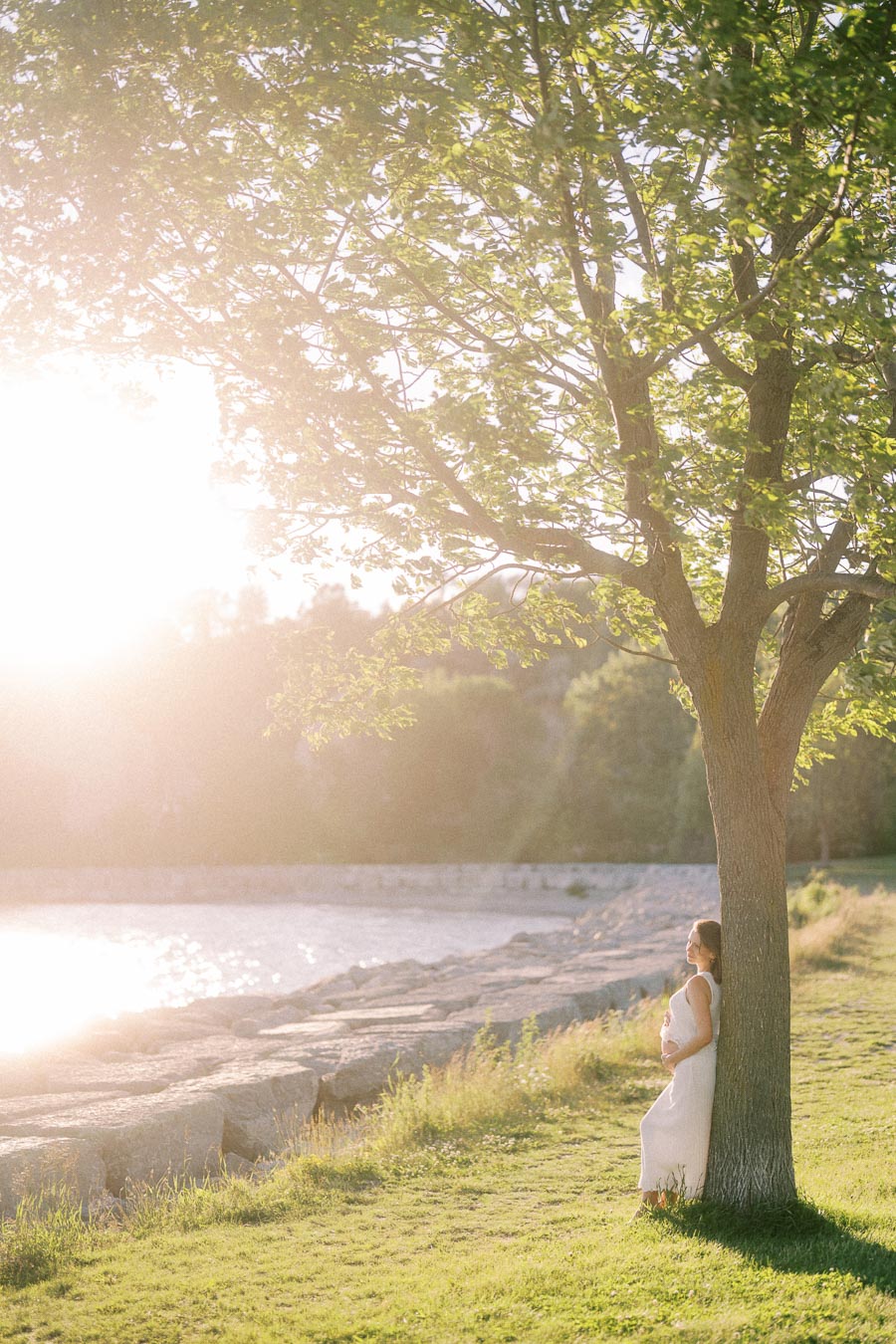 Pregnant woman in a white dress stands beneath a tree beside the sunlit Lake Ontario shoreline.