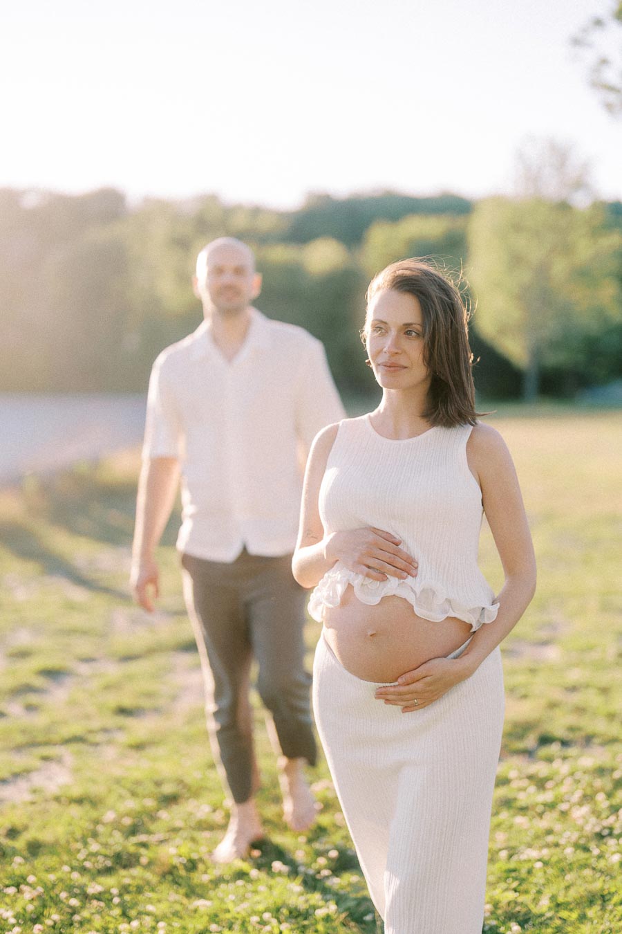 Pregnant woman in a white outfit walks through a grassy park with her partner behind her.