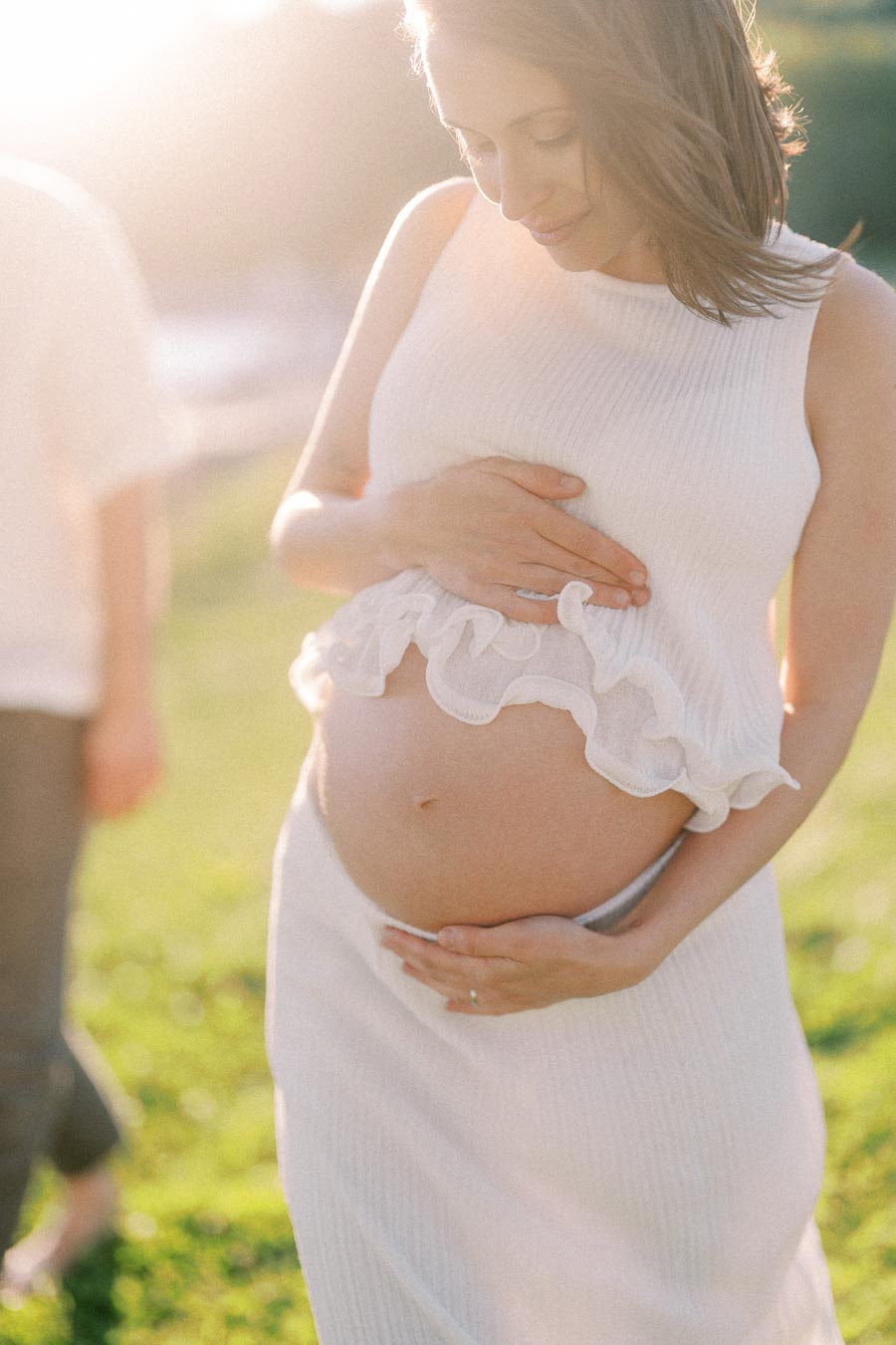 Pregnant woman in a white dress cradles her belly in warm evening sunlight.