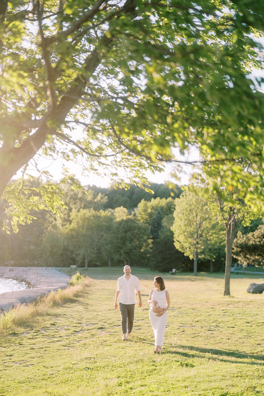 Expectant couple walking through a grassy park beside the water in warm evening light.