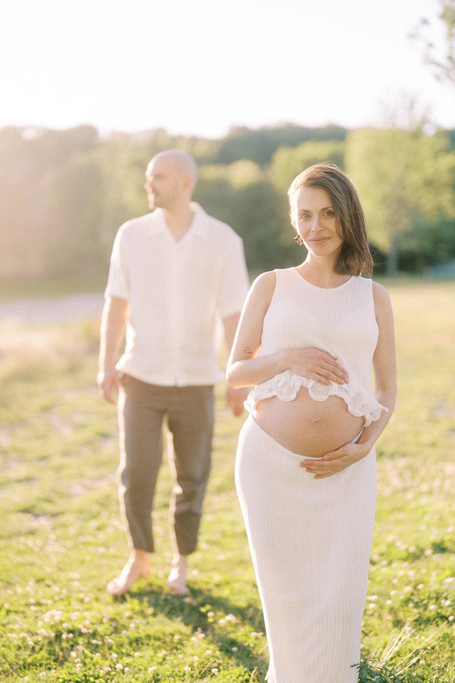 Pregnant woman in a white dress holds her belly while her partner stands behind her in a sunny field.