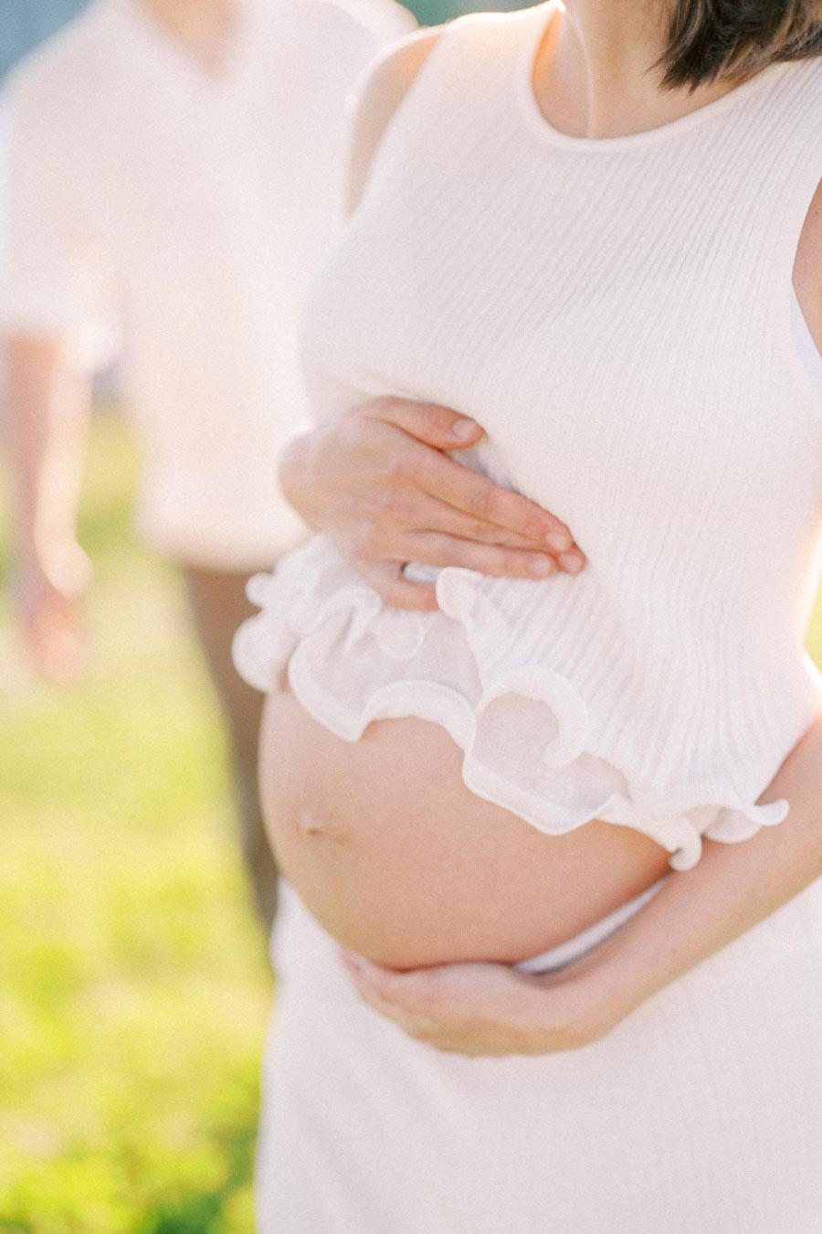 Close-up of a pregnant woman in white holding her belly outdoors in soft sunlight.