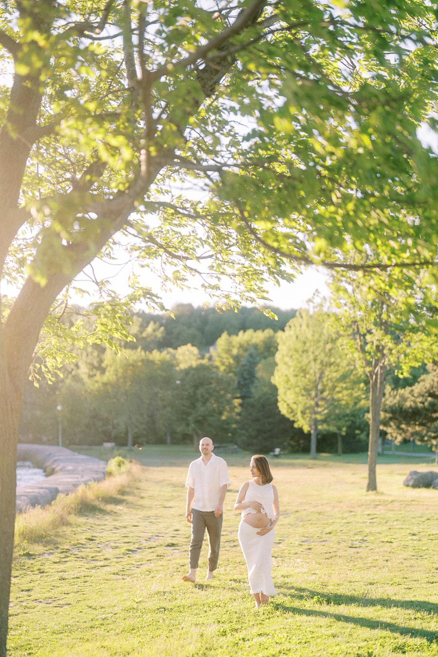 Pregnant woman in a white dress walks with her partner through a sunny grassy park.