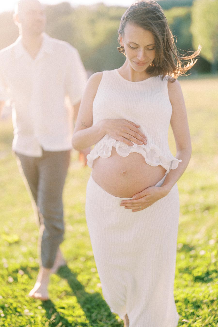 Pregnant woman in a white dress holds her belly while a man walks behind her in sunlight.