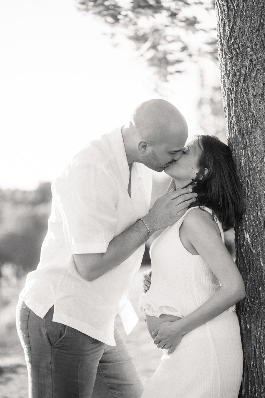 Expectant couple kissing beside a tree, with the woman holding her baby bump.