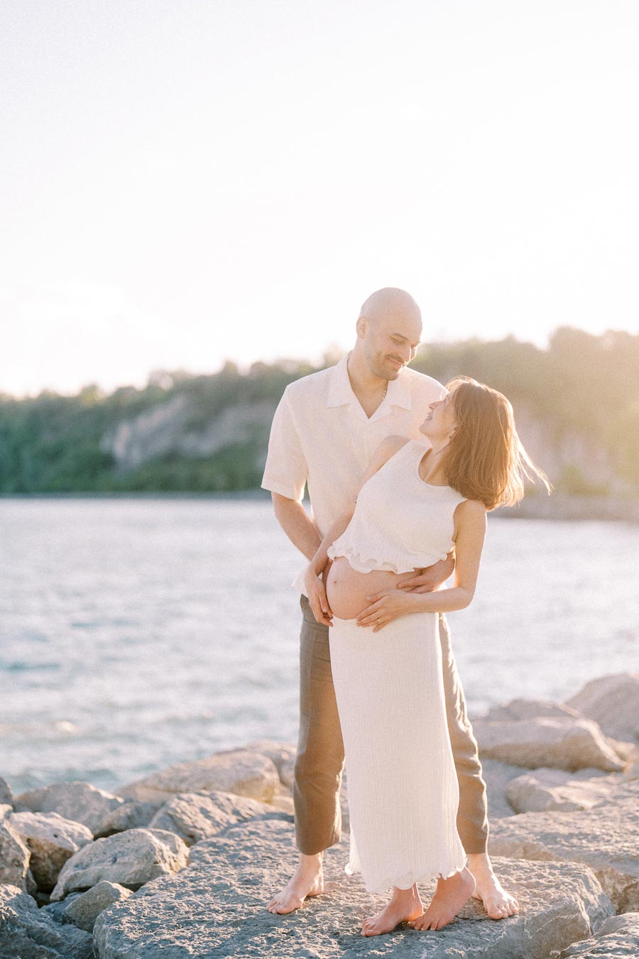 Expectant couple standing barefoot on rocks by the water at Scarborough Bluffs.