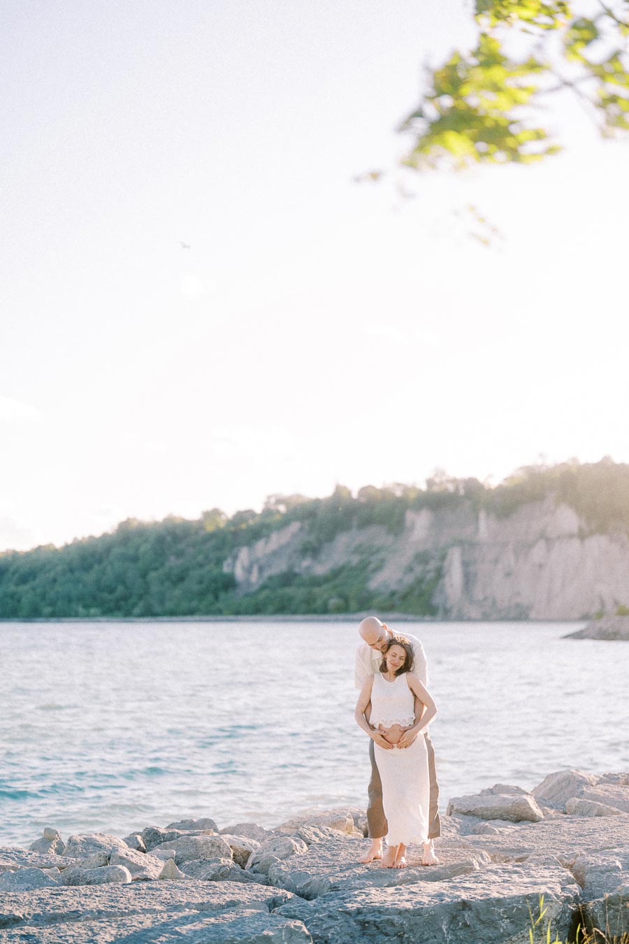 Expectant couple stands on lakeside rocks with Scarborough Bluffs and Lake Ontario behind them.