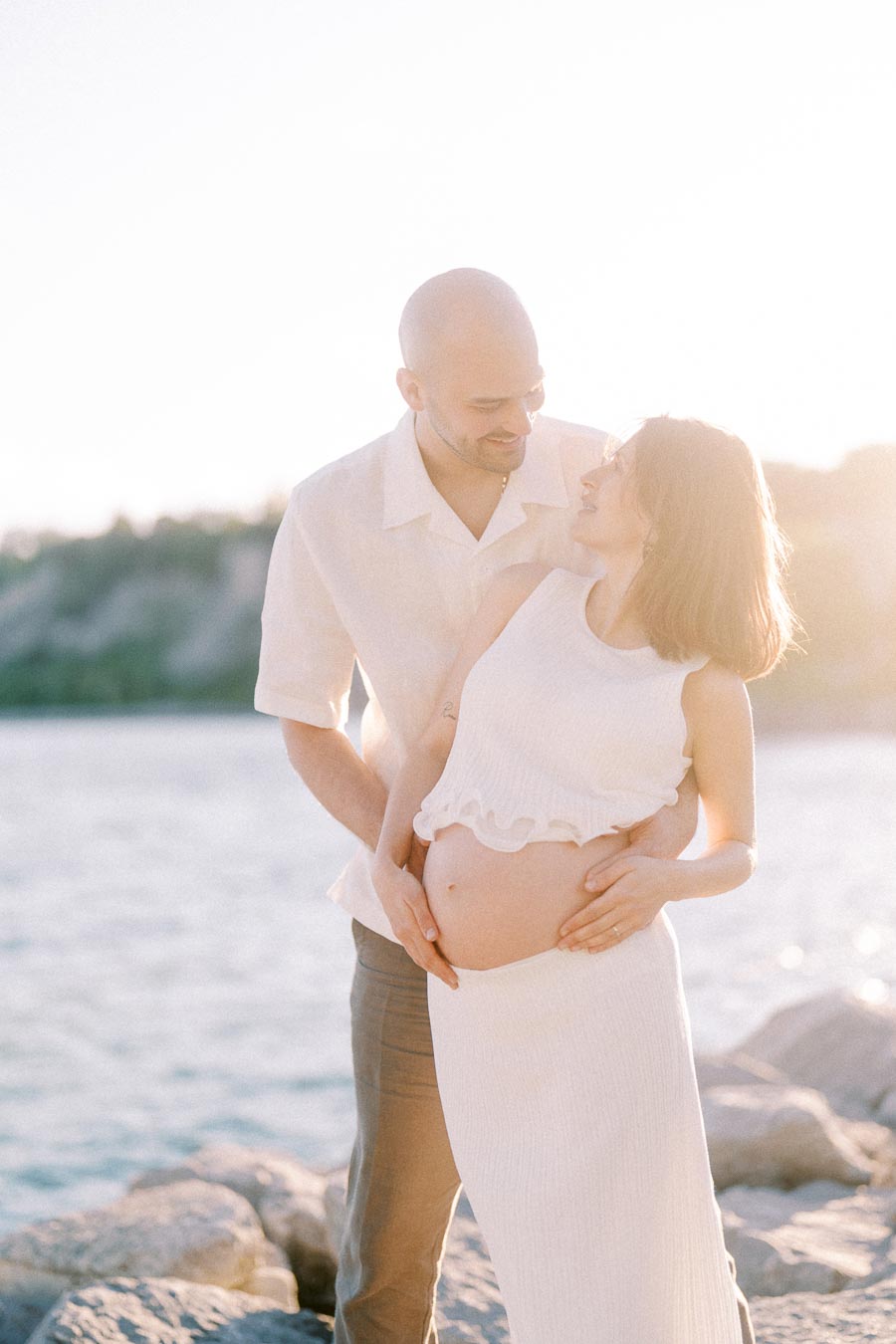 Pregnant woman and partner stand together on rocks by the water in warm backlight.