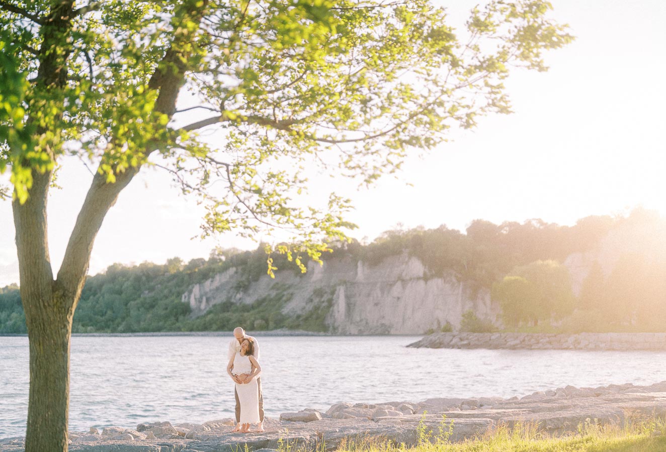 Expectant couple stands on rocky Lake Ontario shoreline under a tree at Scarborough Bluffs.